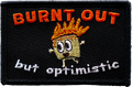 Embroidered patch with cartoon character and text 'BURNT OUT but optimistic' on a black background