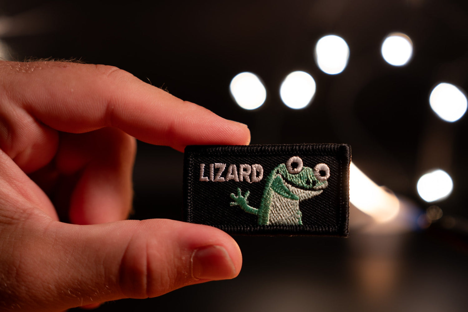 Hand holding a patch with a lizard design and 'Lizard' text against a blurred light background