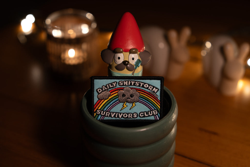Gnome figurine with a 'Daily Shitstorm Survivors Club' patch on a wooden surface with candles in the background.