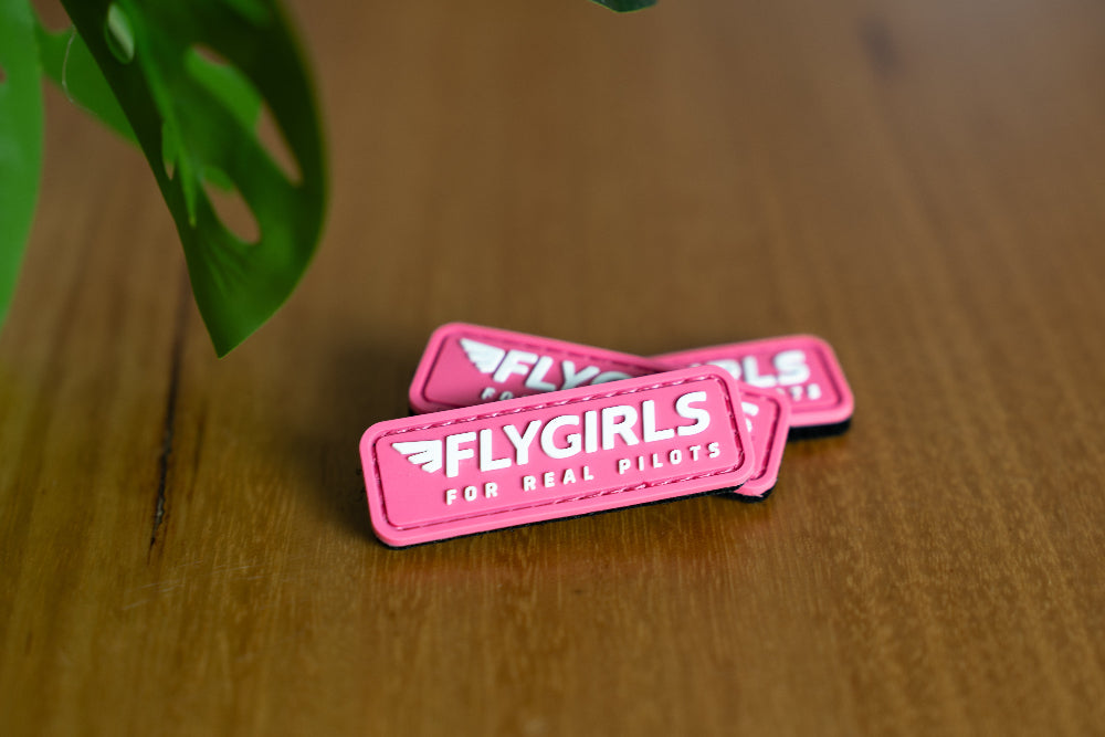 three pink patches with 'FLYGIRLS FOR REAL PILOTS' text on a wooden surface.