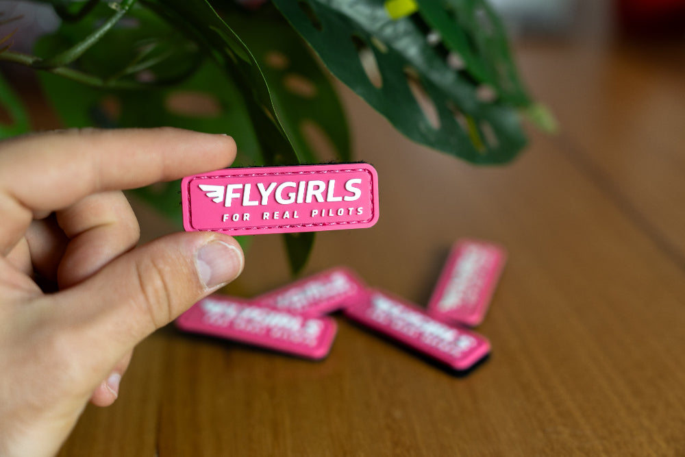 Hand holding a pink 'FLYGIRLS' patch with more patches on a wooden surface.