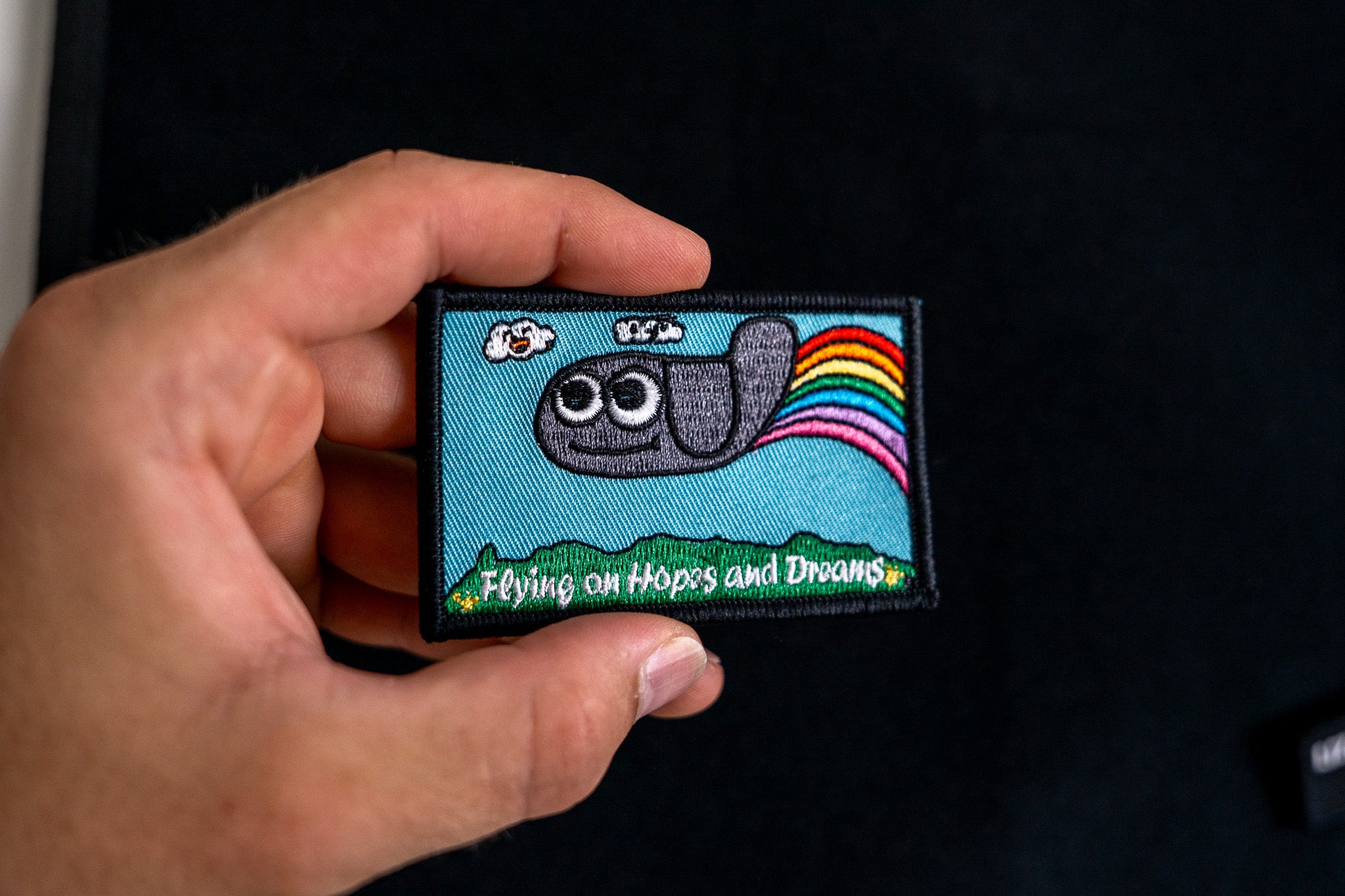 Hand holding a colorful embroidered patch with a cartoon character and rainbow on a dark background