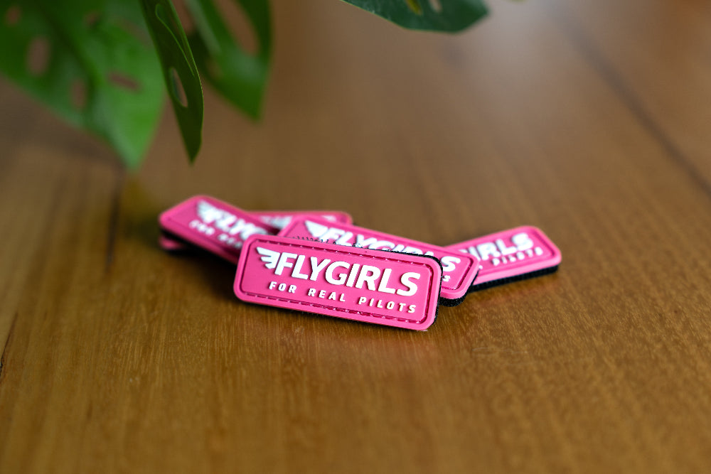 Pink 'FLYGIRLS' patches on a wooden surface with a plant in the background